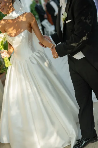 Ceremony moment as bride and groom holding hands, her strapless gown and calla lily bouquet beside a black tuxedo, on an outdoor aisle with white chairs
