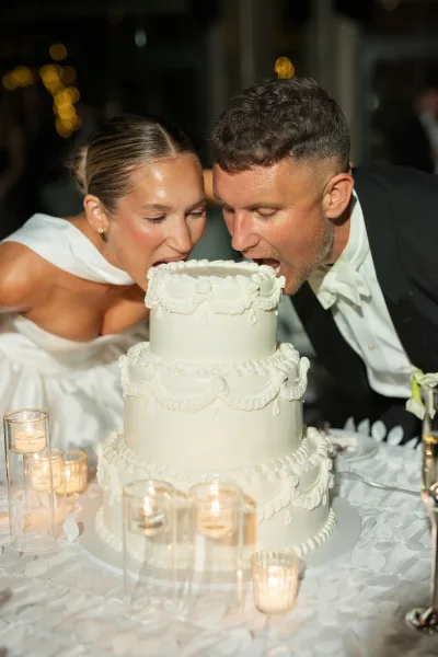 Wedding cake cutting as bride and groom share a cake bite beside a three-tier piped cake on a candlelit table with bokeh lights