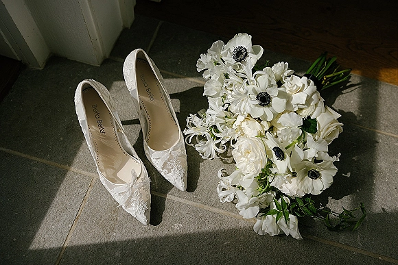 Bridal shoes with lace wedding heels beside a white anemone, rose, and sweet pea bouquet on stone tile in soft window light