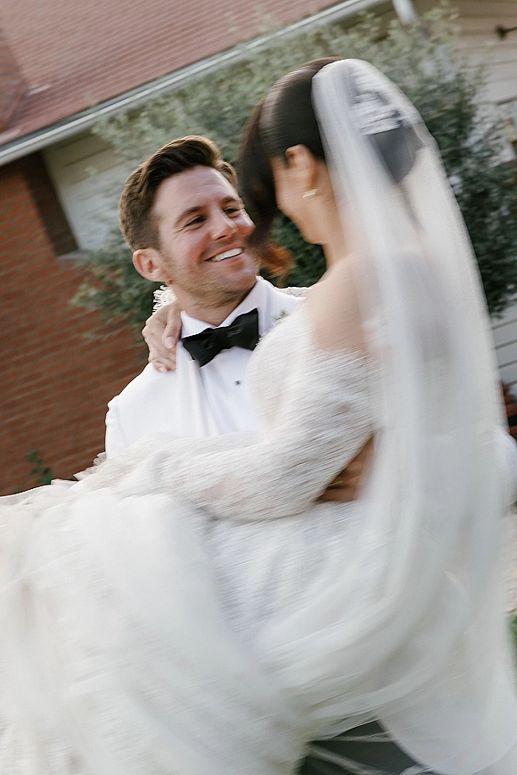 Couple portrait of groom holding bride in a wedding dip pose, her lace strapless gown and veil flowing before a brick wall and greenery