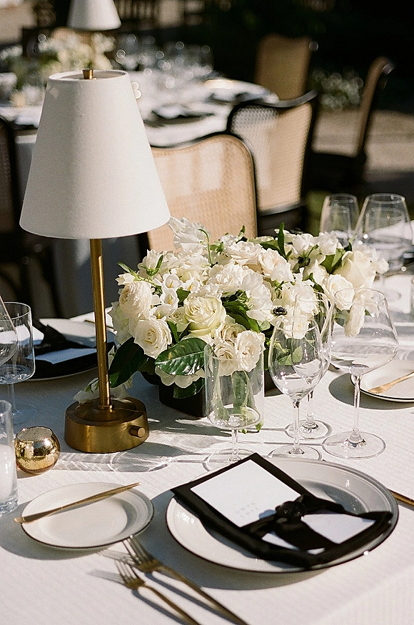 Reception tablescape with white wedding tablescape details, white rose centerpiece, brass table lamp, gold flatware, and black ribbon place cards