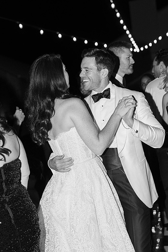 First dance as bride and groom dancing close, her strapless lace wedding dress and his tuxedo under string lights in a dark reception space