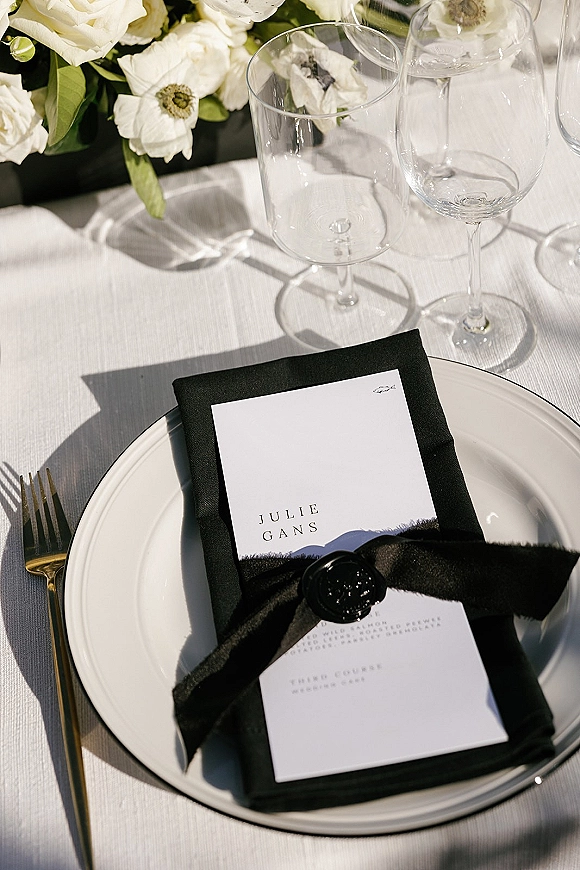 Reception tablescape with a wedding place setting featuring menu and place cards tied with black ribbon and wax seal, gold flatware, wine glasses