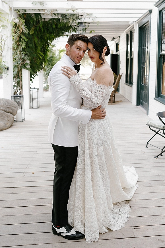 Couple portrait of bride and groom embrace on a covered porch, her lace wedding dress train beside his white tuxedo jacket under hanging greenery