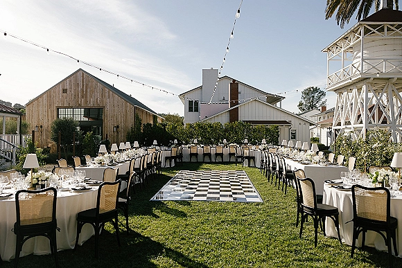 Outdoor reception setup with long banquet tables in white linens, cane-back chairs, floral centerpieces and string lights by a barn lawn