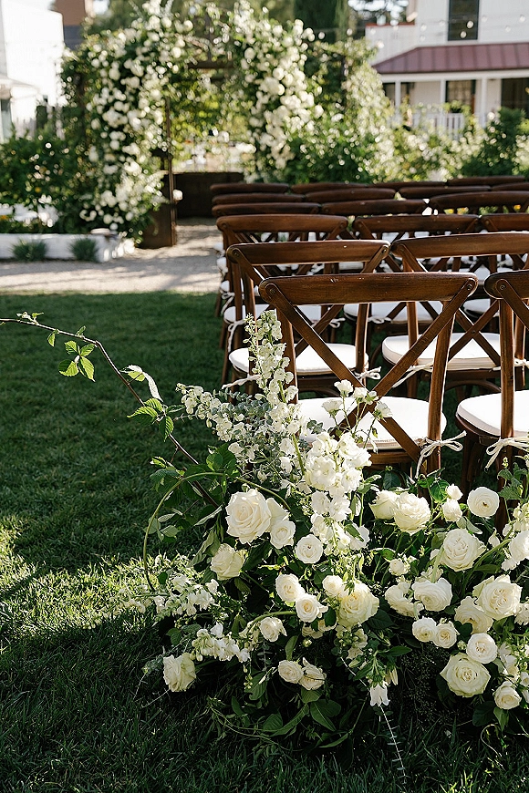 Ceremony setup with outdoor wedding ceremony seating facing a floral arch of white roses and greenery on a lawn beside a house