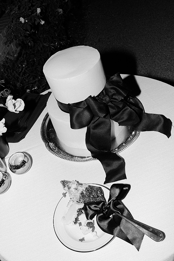 Wedding cake with white fondant cake finish and a black satin ribbon bow on a stand, styled with candles, knife and plate on white linen tablecloth