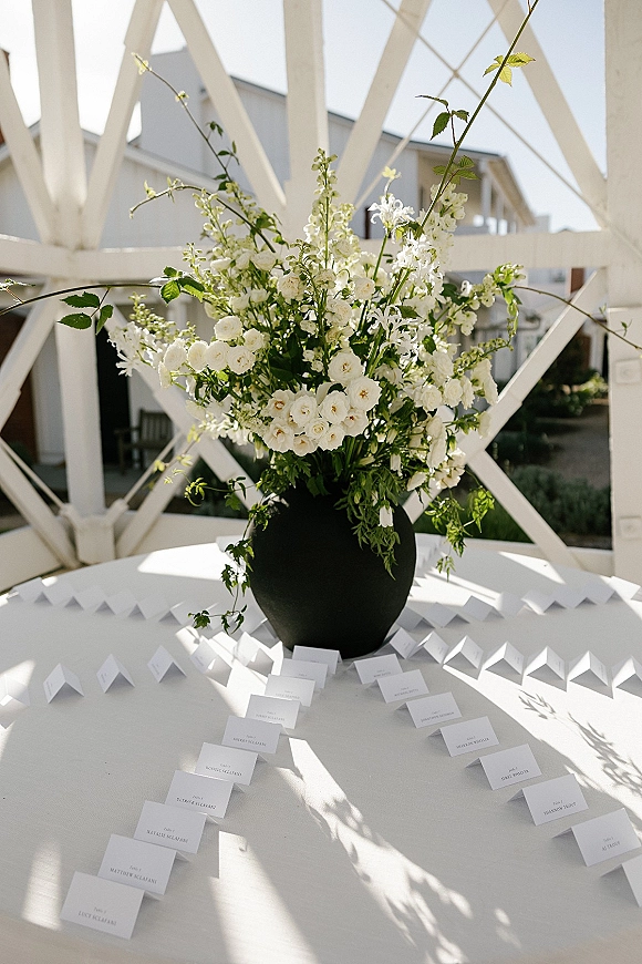 Wedding escort cards arranged on a round display table with folded place cards, a black vase, and white flowers with greenery outdoors in daylight