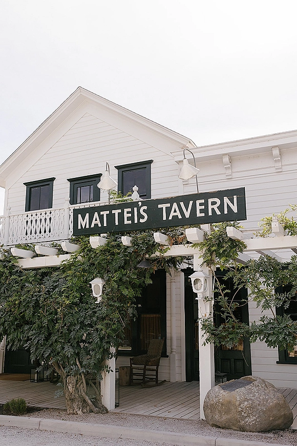 Wedding venue exterior with a hanging sign at a vine-covered pergola entrance, white clapboard building with black shutters and porch behind