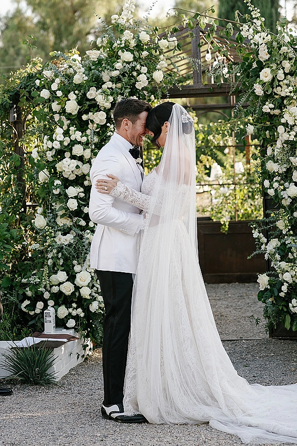 Wedding couple portrait of bride and groom embrace with forehead touch under a white rose floral arch, hanging lights and greenhouse backdrop