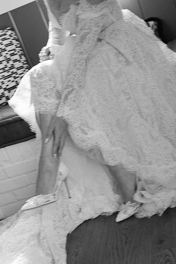 Wedding dress close-up with lace wedding dress long sleeves and train, bridal heels peeking out beside a couch and brick fireplace