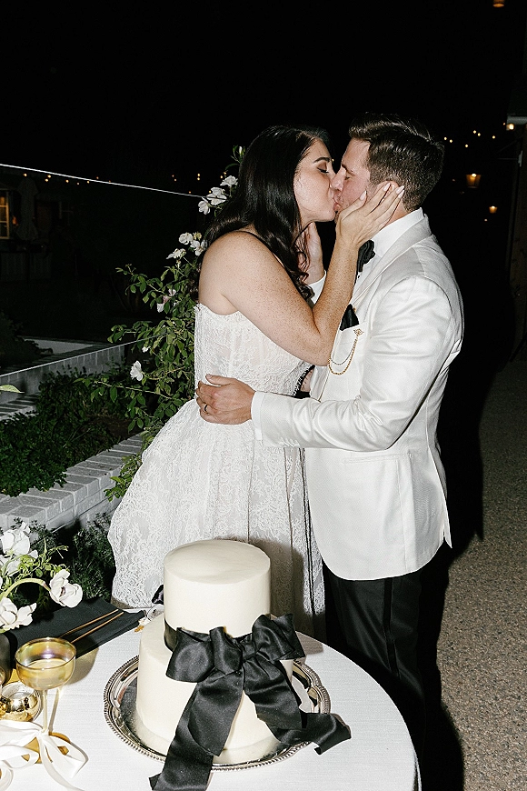 Wedding kiss beside the cake cutting moment, bride in lace dress and groom in white tux under string lights on a garden patio at night