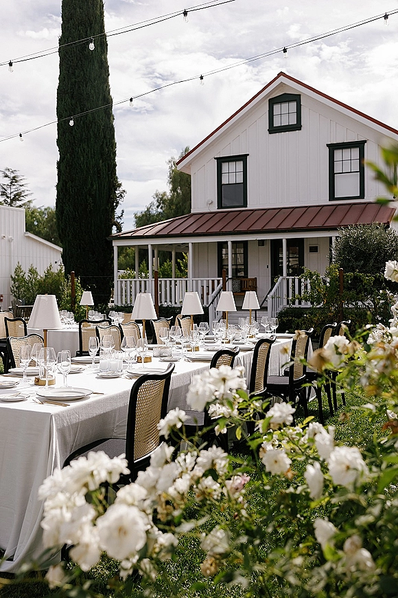 Outdoor reception tablescape with garden reception tables, long white linens, rattan bistro chairs, and string lights by a white farmhouse porch