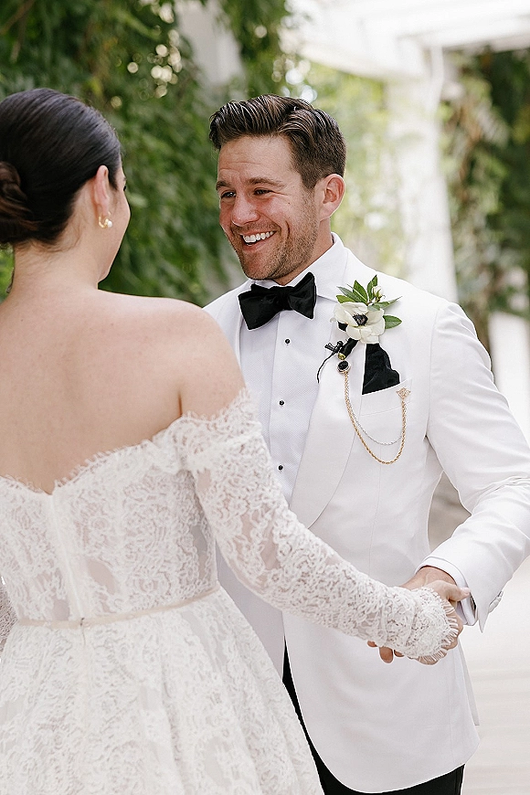 First look moment as bride in an off-the-shoulder lace gown holds hands with groom in white tux on a greenery walkway by columns