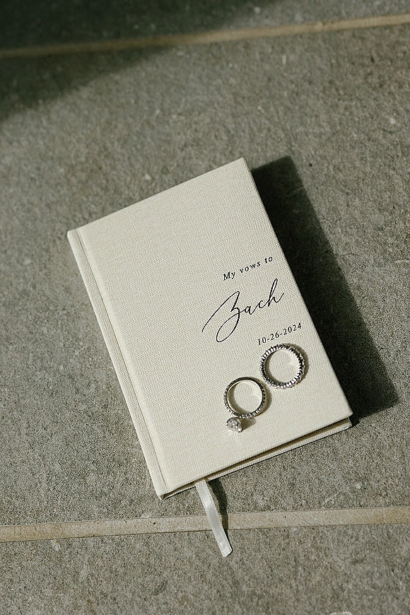 Wedding vow book flat lay with engagement ring and wedding band beside a ribbon-bookmarked vow booklet on stone tile floor