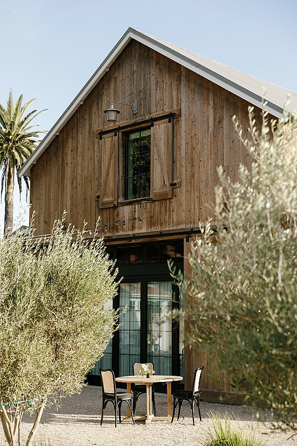 Barn wedding venue with rustic barn exterior, glass double doors and bistro patio seating on gravel, framed by olive trees under blue sky