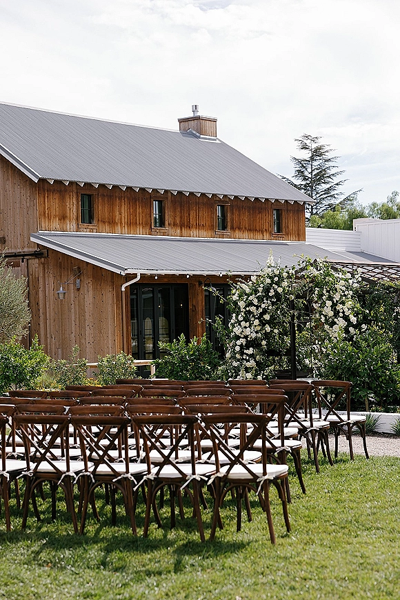 Ceremony setup with outdoor wedding ceremony seating, wood chairs and white cushions facing a floral arch under string lights by a barn