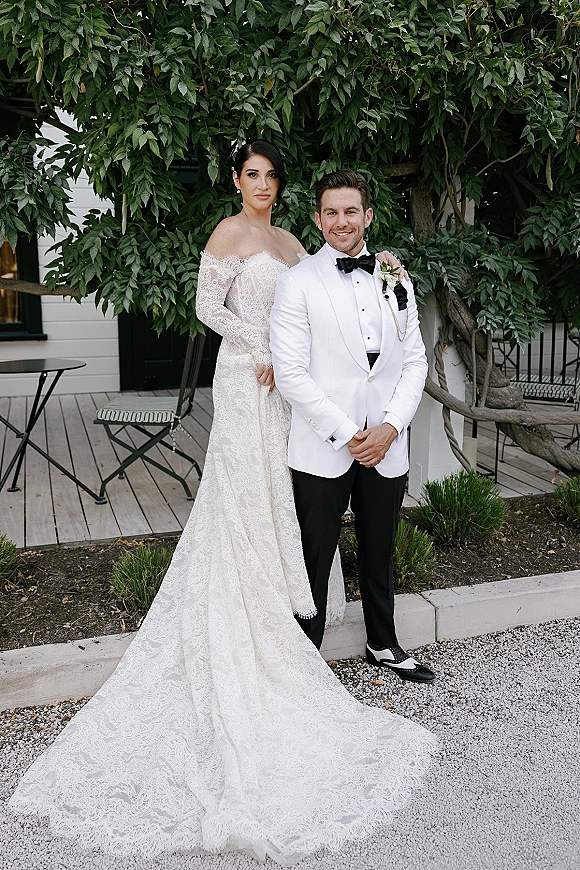 Couple portrait of bride in off-the-shoulder lace gown and groom in white tux jacket, posing on a leafy patio by a white wall