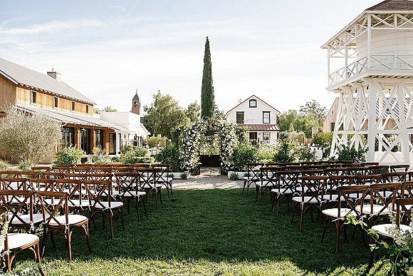 Outdoor ceremony setup with garden wedding ceremony seating, crossback chairs lining an aisle to a floral arch on a grassy courtyard lawn