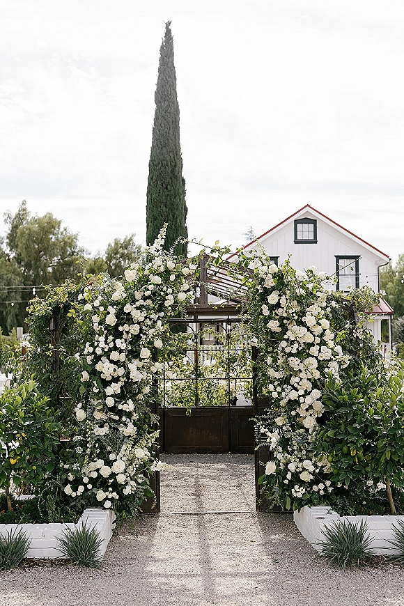 Wedding entrance arch with white rose and greenery garland on a wooden gate, string lights above a gravel path to a farmhouse