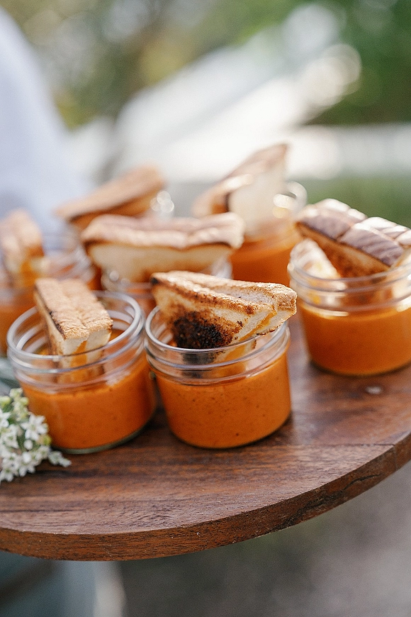 Wedding appetizers of tomato soup in mason jars with mini grilled cheese bites on a wooden board, set against outdoor greenery in natural light