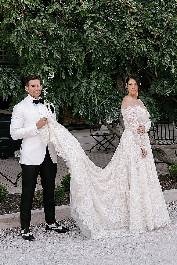 Couple portrait of bride in an off-the-shoulder lace gown with long train as groom in white dinner jacket holds it on leafy patio
