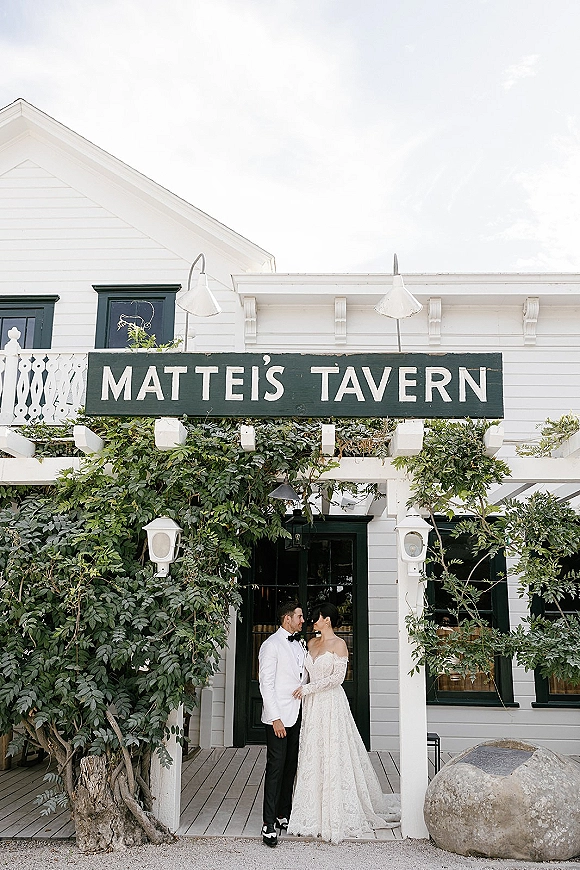 Couple portrait of bride in a wedding dress and groom in a tuxedo gazing at each other by an ivy-covered white clapboard porch entrance.