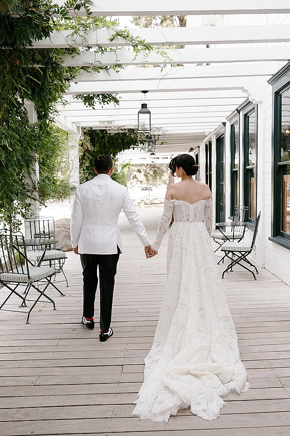 Couple portrait of bride and groom walking away holding hands, her off-shoulder lace dress train trailing under vine-covered pergola lights