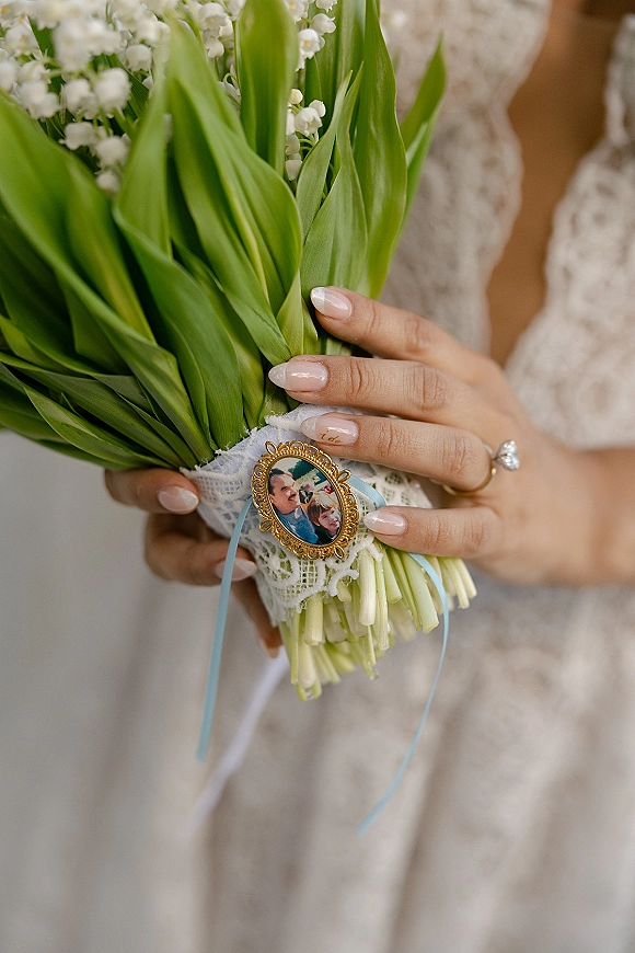 Wedding bouquet of lily of the valley held in a lace-wrapped handle with blue ribbon and photo locket charm against a neutral indoor backdrop