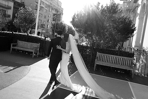Couple portrait of bride holding groom’s arm as they walk away down a sunlit city street, her long cathedral veil trailing behind