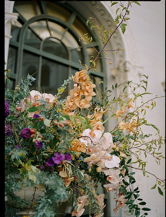 Wedding floral arrangement with orchid wedding flowers, peach roses, greenery and branches in an urn by an arched window on stone facade