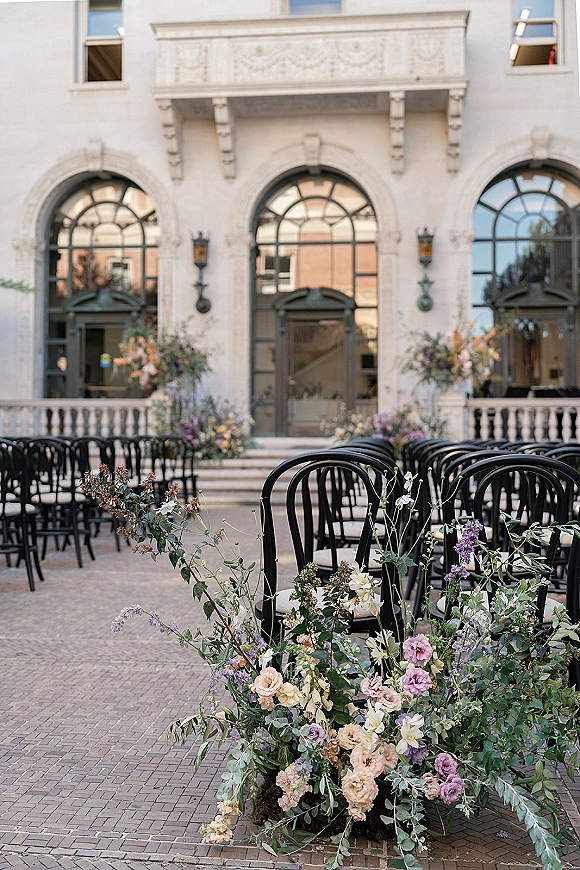 Ceremony setup with outdoor ceremony seating and black chairs lining a floral aisle, pastel arrangements on stone steps before a white arched facade