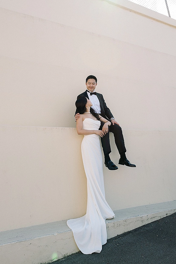 Couple portrait of bride and groom pose, her strapless dress train draped as he sits on a concrete ledge by a neutral wall