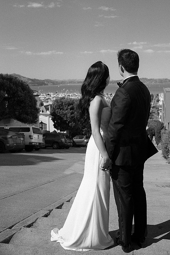 Couple portrait of bride and groom from behind holding hands, her strapless wedding dress train and his tuxedo by a bay city overlook