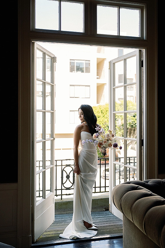 Bridal portrait of a bride holding bouquet in a strapless satin gown, looking over her shoulder by open French doors with city view