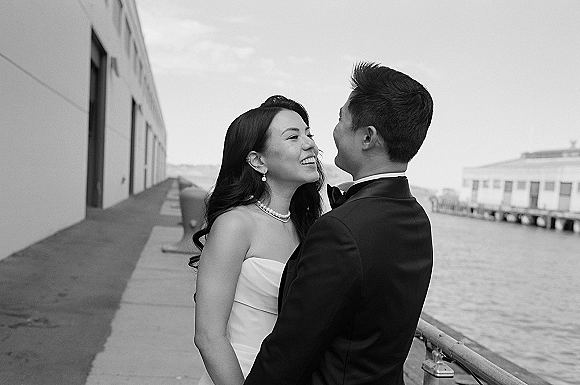 Couple portrait of bride and groom laughing face to face, holding each other on a waterfront pier with industrial buildings behind them