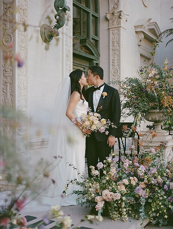 Wedding kiss portrait of bride and groom kissing on stone steps, bride in strapless dress with veil holding bouquet before ornate facade