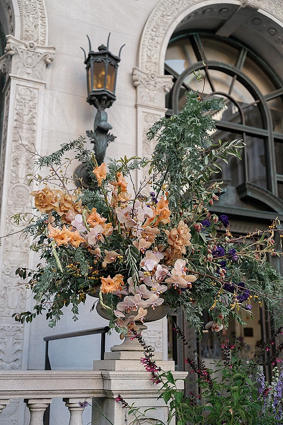 Wedding floral arrangement in a stone urn with orchids, peach blooms, and greenery against an ornate stone facade with arched window
