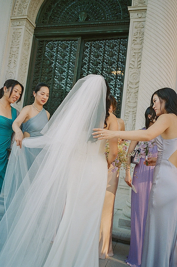 Bride with bridesmaids fixing veil as she stands on stone steps by an ornate wrought iron doorway, holding a bouquet in a strapless gown