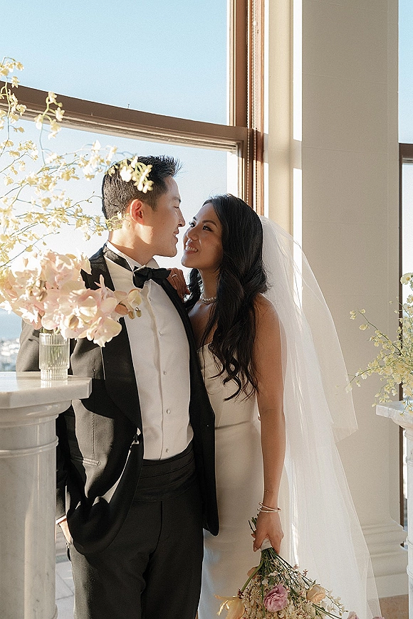 Couple portrait of bride and groom posing by sunlit windows, gazing at each other as she holds an orchid bouquet with veil accent