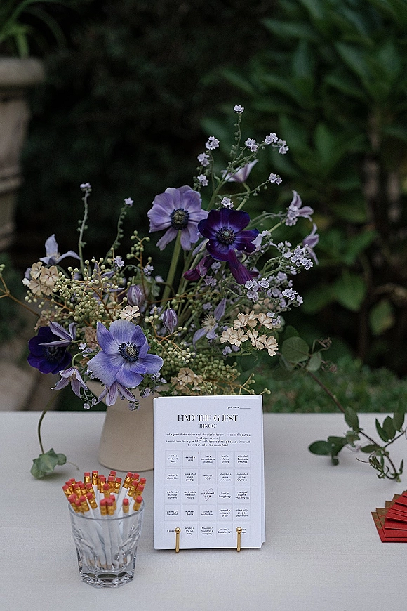 Wedding guest book table with guest book bingo cards, pencils, and a wildflower arrangement in a vase against outdoor greenery backdrop
