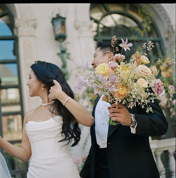 Couple portrait of bride and groom laughing, groom holding bouquet by a stone building with arched windows and vine greenery