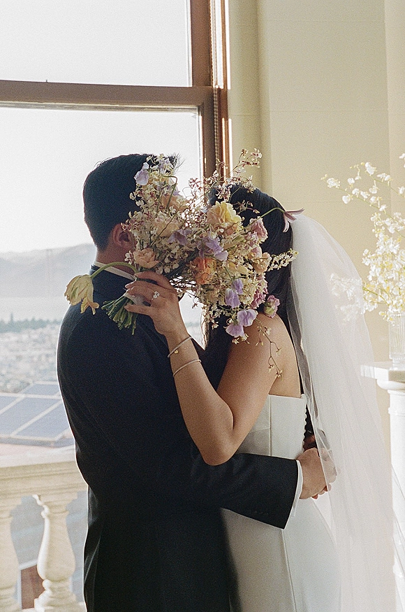 Wedding kiss portrait of bride and groom kissing behind a wildflower bouquet, veil and tux framed by sunlit window with city view