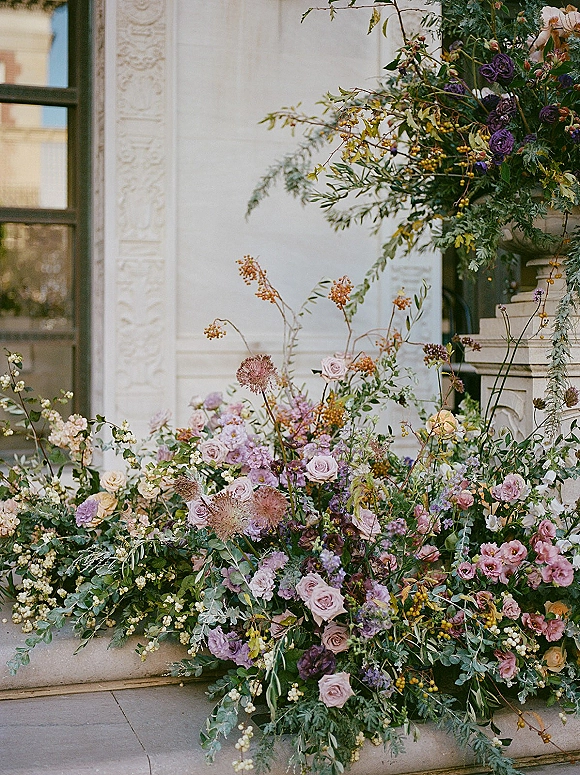 Wedding floral installation of roses, wildflowers, and greenery in an urn planter on stone steps beside an ornate doorway with window