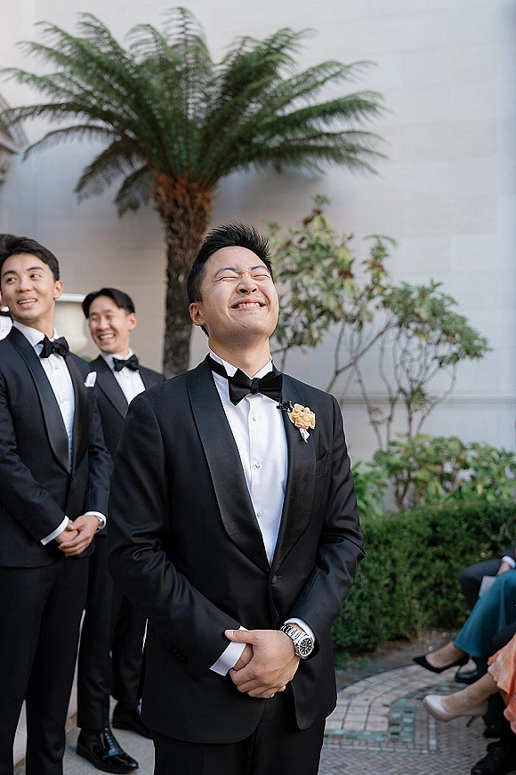 Groom portrait in a black tuxedo with bow tie and boutonniere, smiling on an outdoor walkway with palm tree and seated guests behind
