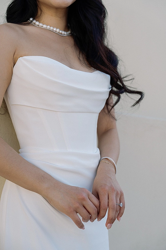 Wedding dress with strapless satin bodice, bride’s hands showing engagement ring and bracelet, pearl necklace against a neutral wall