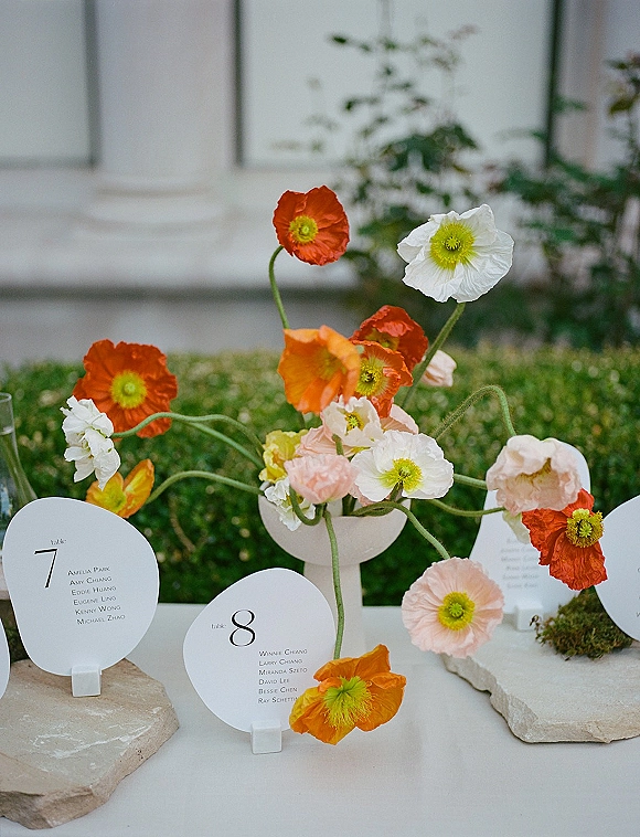 Wedding escort cards with round escort cards on stone holders, set beside a ceramic bud vase with poppy flowers on moss in a garden setting