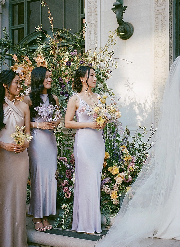 Bridesmaids in lavender dresses holding pastel bouquets, lined up on stone steps by a carved facade with a window and sconce