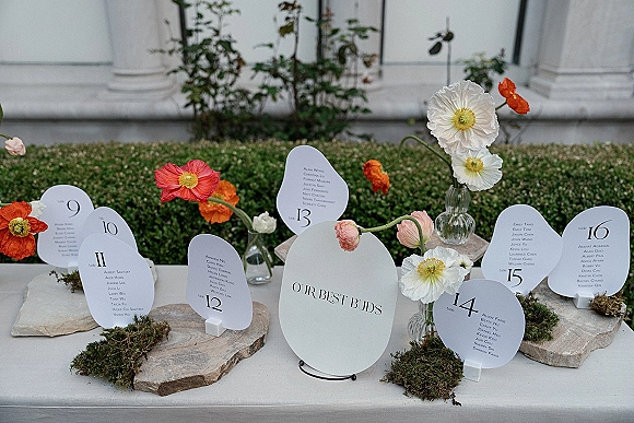 Wedding escort cards arranged on stone slabs with moss accents, table numbers, and poppy bud vases on a linen table by hedges and columns