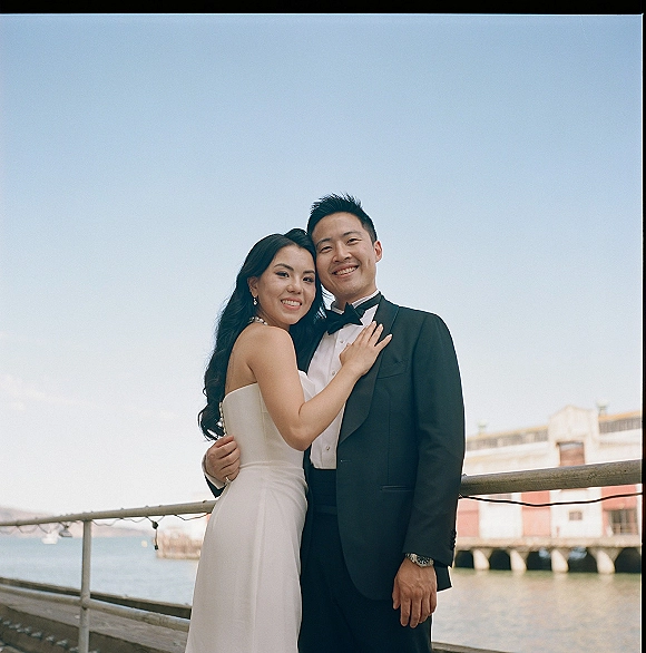 Couple portrait of bride hugging groom in tuxedo and strapless dress, pearl necklace accent, smiling by waterfront pier railing with string lights